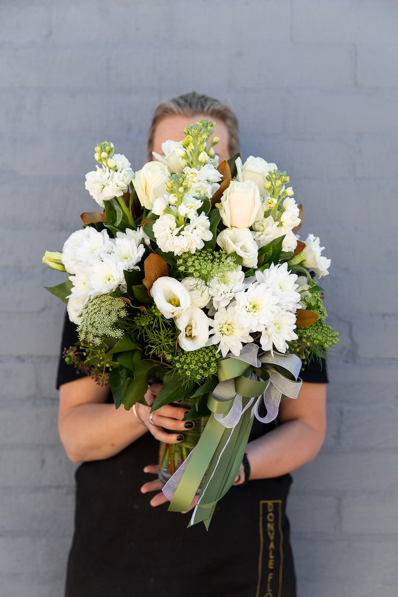 A Donvale Flower Gallery team member holding the Alaska bouquet which consists of cream and white blooms with lush foliage and presented in a glass cylinder vase.