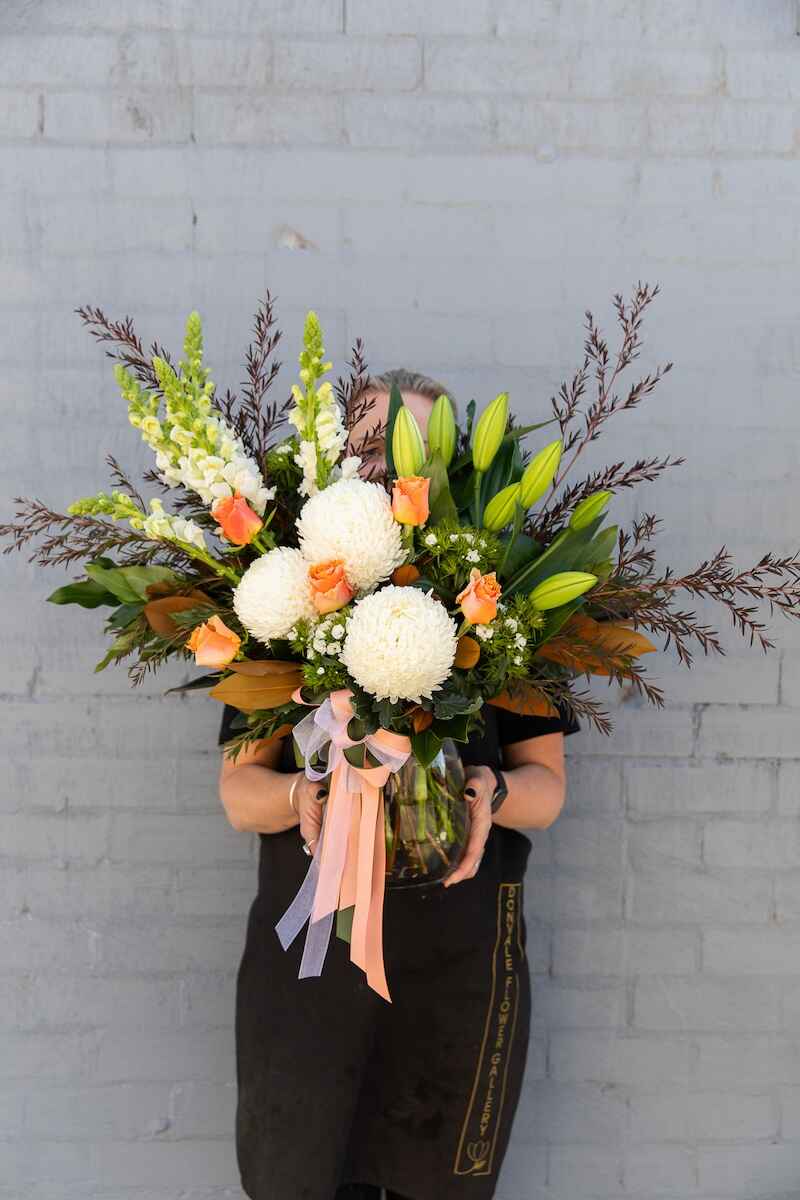 Photo of a Donvale Flower Gallery team member holding Christobelle - a modern-style vase arrangement with white lilies, apricot roses and snapdragons. Comes with a complementary bow.