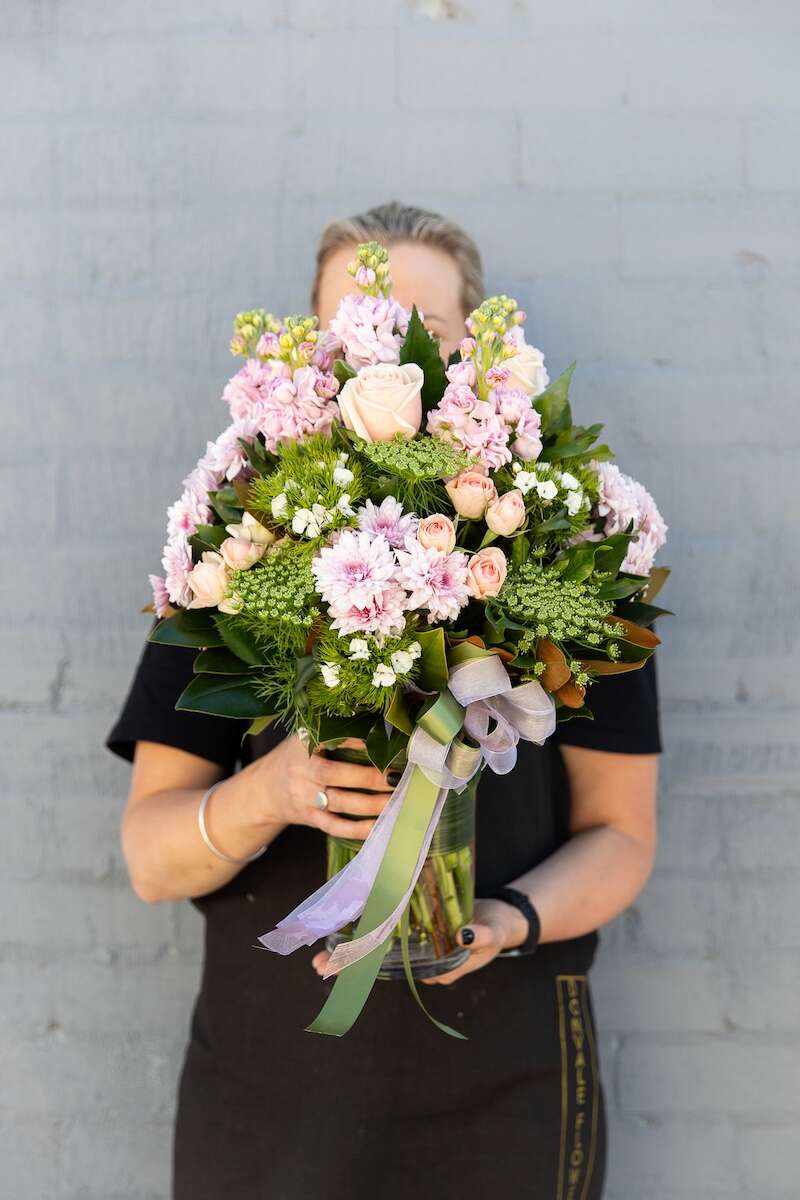 Image of a Donvale Flower Gallery team member holding the Delightful flower bouquet - consisting of pink and white blooms with lush foliage. Comes in a leaf-lined glass vase.